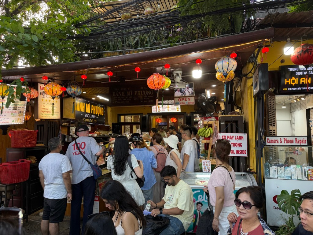 A busy street scene in Hoi An, showcasing the famous Banh Mi Phuong restaurant with colorful lanterns and a crowd of people waiting to order.